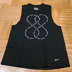 Svart Nike Dri-Fit linnetopp XS - Svart linnetopp från Nike i Dri-Fit material, perfekt för träning. Framsidan har ett stort tryck med texten 'Nike Training Club' i en cool, cirkulär design. Toppen är ärmlös och har en loose passform för extra rörelsefrihet.