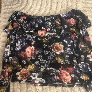 Blommig offshoulder topp från H&M - Supersnygg offshoulder topp från H&M med blommigt mönster i mörkblått, rosa, lila och grönt. Toppen har volangdetalj längs halsen och är långärmad. Perfekt för dig som gillar romantiska vibes och vill sticka ut med färg och mönster.