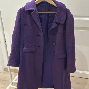 Lila kappa från Martinette - Snygg lila kappa från Martinette med klassisk krage och två knappar framtill. Jackan har två fickor med lock och är helfodrad i matchande lila tyg. Perfekt för dig som vill sticka ut med färg i höst och vinter.