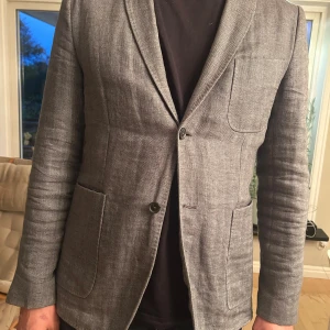 Grå kavaj från J.Lindeberg skinny fit - Snygg grå kavaj från J.Lindeberg i skinny fit-modell. Kavajen har två knappar framtill, tre utanpåliggande fickor och klassisk krage. Materialet är linne och ger en lätt och stilren känsla. Perfekt för dig som gillar en smalare passform och modern look.