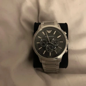 Emporio Armani silverfärgad klocka - Stilren klocka från Emporio Armani med silverfärgat armband i rostfritt stål och svart urtavla. Klockan har kronograf-funktion, datumvisning och klassisk rund form. Snygga detaljer med logga på både urtavla och spänne. Perfekt accessoar för dig som gillar en clean look.