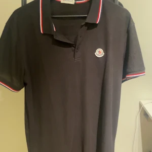 Svart pikétröja från Moncler - Svart pikétröja från Moncler med klassisk krage och korta ärmar. Tröjan har Moncler-logga på bröstet och snygga rödvita ränder på krage och ärmslut. Tillverkad i mjuk bomull som känns skön mot huden. Perfekt för dig som gillar stilrena och sportiga plagg. Tröjan är i XL men sitter som L