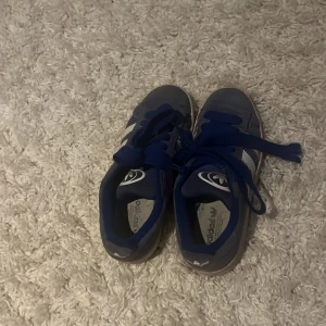 Adidas Campus blå sneakers - Säljer ett par Adidas Campus sneakers i blå mocka med vita detaljer och klassiska tre ränder på sidan. Skorna har blå snörning, vit sula och Adidas logga på hälen. Perfekta för dig som gillar retrostil och streetwear.