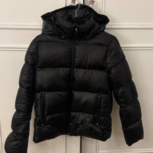 Svart pufferjacka från Calvin Klein Jeans - Svart pufferjacka från Calvin Klein Jeans med huva och två sidofickor med dragkedja. Jackan har en stilren look med breda, quiltade sektioner och hel dragkedja framtill. Perfekt för kalla dagar och enkel att matcha med din streetstyle.