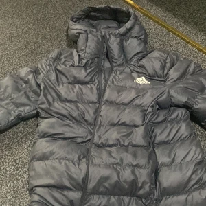 Svart pufferjacka från Adidas, L - Svart pufferjacka från Adidas i storlek L. Jackan har huva, dragkedja framtill och Adidas-logga på bröstet. Den är quiltad och har en klassisk, sportig look som passar perfekt när det är kallt ute. Materialet är syntet och jackan är långärmad. Perfekt för kallare dagar