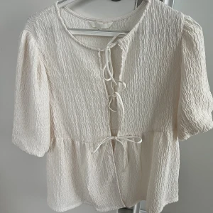 Vit blus med snörning och struktur - Säljer en vit blus med snygg struktur i tyget och tre snörningar framtill. Blusen har korta ärmar och en lätt volang nedtill. Perfekt för dig som gillar romantisk och trendig stil. Superfin till jeans eller kjol! 