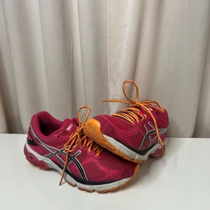 ASICS Gel-Hurako rosa sneakers - Säljer ett par ASICS Gel-Hurako sneakers i en snygg rosa färg med orange snören och detaljer. Skorna har en rund tå, platt sula och är tillverkade i ventilerande syntetmaterial med svarta och vita mönster längs sidorna. Perfekta för träning eller promenader.