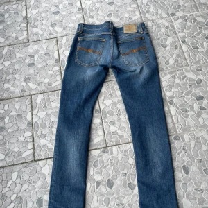 Blå  jeans från Nudie Jeans - Säljer ett par klassiska blå  jeans från Nudie Jeans med snygga slitningar och orangea sömmar på bakfickorna. Jeansen har fem fickor, dragkedja och är tillverkade i mjukt denimtyg. Perfekt passform och stilren look.
