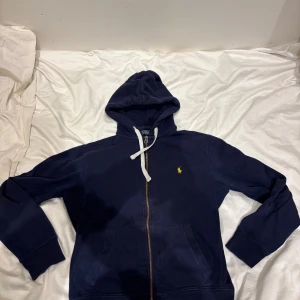 Marinblå hoodie från Polo Ralph Lauren - Snygg marinblå hoodie från Polo Ralph Lauren med gul broderad logga på bröstet. Tröjan har huva med vita snören, dragkedja framtill och två fickor. Tillverkad i mjuk bomull, perfekt för chill dagar. Klassisk och stilren design som alltid funkar i vardagen