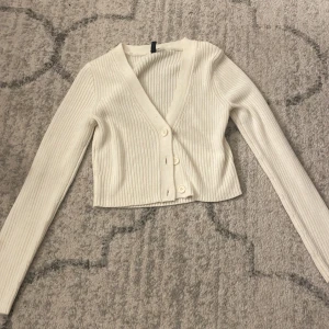 Ljusbeige ribbad kofta med v-ringning - Säljer en ljusbeige croppad kofta med ribbad struktur och v-ringad hals. Koftan har långa ärmar och tre knappar framtill. Perfekt för dig som gillar stilrena och enkla plagg med snygg passform.