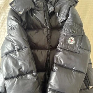 Svart pufferjacka - Svart pufferjacka med glansig finish. Jackan har dragkedja framtill och hög krage med tryckknappar. Perfekt för kalla dagar. Säljs då den inte kommer till användning. OBS: dragkedjan går oftast inte att stänga, men bör lätt kunna fixas. Jättebra kvalité annars och otroligt varm att ha! Kan skickas inom 1-2 dagar. 