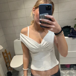 Vit topp från Zara - Supersnygg vit omlottopp från Zara med offshoulder och lätt rynkade detaljer vid bysten. Toppen har smala axelband och sitter figurnära. Perfekt för dig som gillar en stilren och trendig look.