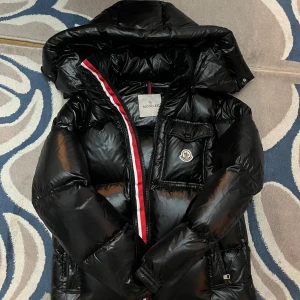 Svart Moncler pufferjacka med huva - Storlek S Svart pufferjacka från Moncler med glansig finish, stor huva och ikonisk logga på bröstfickan. Jackan har dragkedja med röd och vit kant, praktiska fickor och quiltad design. Perfekt för kalla dagar när du vill ha både stil och värme.