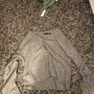 Grå kofta från 157 med knappar - Mysig grå kofta från 157 i storlek S. Koftan har två knappar framtill, lång ärm och en enkel, stilren design. Perfekt att slänga på sig över en t-shirt eller linne. Materialet är mjukt och känns skönt mot huden.