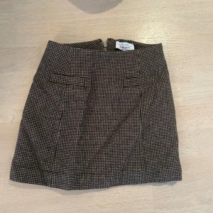 Rutig brun minikjol från Aéryne - Snygg brun och svart rutig minikjol från Aéryne med klassiskt mönster och diskreta fickdetaljer framtill. Kjolen har en rak passform och dragkedja bak, perfekt till stickade tröjor eller boots.