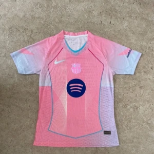 Barça x Nike rosa fotbollströja - Säljer en Barcelona fotbollströja från Nike i rosa och ljusblå toner. Tröjan har korta ärmar, meshmaterial och tryck med Spotify-logga på bröstet samt UNHCR ACNUR på ryggen. Snygga grafiska linjer och klubbmärke framtill. Perfekt för dig som älskar unik design.