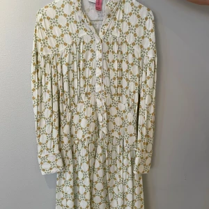 Blommig kortklänning från Sandro Paris - Superfin kortklänning från Sandro Paris med blommigt mönster i vitt, grönt och gult. Klänningen har långärmade ärmar, knappar framtill och en mjuk, flowy passform. Materialet är bomull och viskos, perfekt för vår och sommar. Snygg krage och fina detaljer vid axlar och midja.