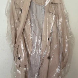 Beige trenchcoat från S.Oliver - Snygg beige trenchcoat från S.Oliver med klassisk dubbelknäppning, axelklaffar och bälte i midjan. Jackan har långa ärmar, randigt foder och detaljerade knappar. Perfekt för dig som gillar stilrena och tidlösa plagg. Bältet i midjan framhäver silhuetten, medan sprundet baktill ger rörelsefrihet. Perfekt för både vardag och fest.  Oanvänd, paketerad i plastenballage  Ordinarie pris ca 1600kr