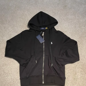Ralph lauren kofta - Helt ny Ralph Lauren zip-hoodie i svart, med tag kvar. Klassisk design med diskret logga på bröstet och högkvalitativa detaljer som metalländar på snören och robust dragkedja. Tillverkad i mjukt och skönt material som ger en perfekt passform och komfort. En stilren och mångsidig kofta som passar till allt – både till vardag och en mer uppklädd look.