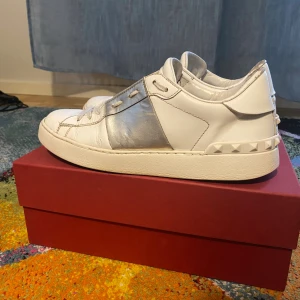 Valentino Garavani sneakers - Snygga Valentino Garavani sneakers i vitt skinn med bred silverdetalj över mitten och ikoniska nitar runt hälen. Klassisk rund tå, platt sula och snörning framtill. Perfekta för dig som vill ha en lyxig och trendig look. Allt finns förutom extra skosnören om ni har några frågor så är de bara skriva
