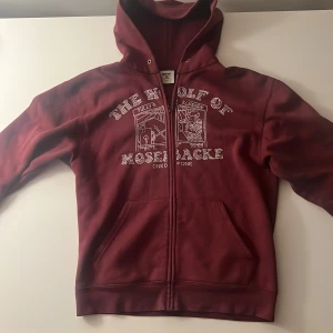 Vinröd hoodie One of One  - Säljer en vinröd hoodie från One of One med huva och dragkedja. Framsidan har ett stort strassmönster med texten 'The Wolf of Mosebacke'. Hoodien har känguruficka och är långärmad, perfekt för dig som vill sticka ut.