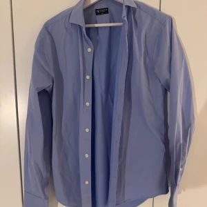Blå skjorta från Tiger of Sweden slim fit - Snygg ljusblå skjorta från Tiger of Sweden med klassisk krage och vita knappar. Skjortan har lång ärm och är tillverkad i mjuk bomull som känns skön mot huden. Perfekt för dig som gillar stilrena och enkla plagg med bra passform.