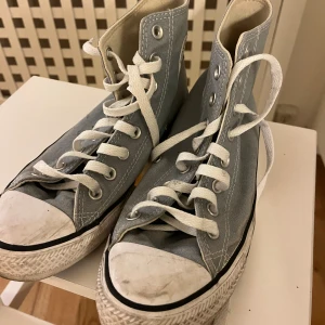 Grå Converse Chuck Taylor All Star - Säljer ett par klassiska Converse Chuck Taylor All Star high-tops i ljusgrått tyg med vita snören och gummisula. Skorna har rund tå, vit tåhätta och tydliga sömmar. Perfekta för dig som gillar en tidlös och avslappnad stil.