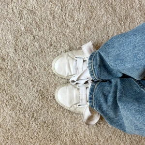 Vita sneakers från Puma med bred snörning - Säljer ett par klassiska vita sneakers från Puma med breda, platta snören och rund tå. Skorna har en stilren design i syntetmaterial och passar perfekt till jeans eller andra avslappnade outfits. Diskret Puma-logga på plösen och sula med bra grepp.