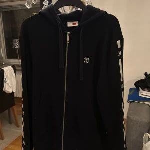 Svart hoodie Tommy Hilfiger x Lewis Hamilton - Svart hoodie från Tommy Hilfiger x Lewis Hamilton med dragkedja och huva. Snygga vita detaljer och H-logga på bröstet och ärmen. Långärmad modell med ribbade muddar och fickor framtill. Perfekt för dig som gillar streetwear och stilrena plagg.
