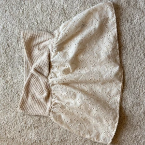 Beige babydoll topp med broderade blommor - Supersöt beige blus med bred, stickad midja och broderade blommor över hela kjolen. Kjolen har en volangig känsla och är gjord i bomull med detaljer i stickat material. Perfekt för dig som gillar romantiska och feminina plagg.