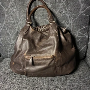 Brun skinnväska från Marc O'Polo - Stilren brun handväska från Marc O'Polo i äkta skinn. Väskan har två rejäla handtag, en praktisk ytterficka med dragkedja och snygga metalldetaljer. Perfekt för dig som vill ha en rymlig och klassisk väska med tidlös design.