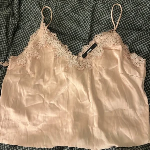 Champagnefärgad linnetopp från Bik Bok - Säljer en champagnefärgad linnetopp från Bik Bok med tunna axelband och vacker spetsdetalj längs v-ringningen. Toppen har en mjuk, glansig yta och är perfekt för dig som gillar romantisk stil. Passar dig som vill ha en feminin och chic look.