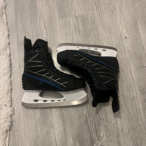 Svarta skridskor Revolution Pulse 5 strl 38 - Säljer ett par svarta skridskor från Revolution, modell Pulse 5, i storlek 38. De har blå och vita detaljer, snörning och en robust design med vit plastskena och stålklinga. Perfekta för dig som vill glida fram på isen med stil.