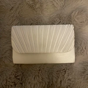 Vit kuvertväska med veckad detalj - Snygg vit kuvertväska med stilren veckad framsida och magnetlås. Insidan har ett fack och en avtagbar silverfärgad kedja. Perfekt storlek för mobil och småsaker. Väskan har en elegant, glansig finish och passar dig som vill sticka ut med en clean look. Defekt: rosa färg på insidan 