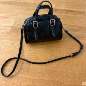 Svart handväska från Zara - Snygg svart handväska från Zara i skinnimitation med både handtag och justerbar axelrem. Väskan har två dekorativa spännen framtill och en dragkedja upptill. Perfekt storlek för det viktigaste och stilren design som funkar till många outfits. 