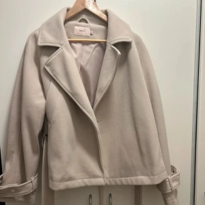 Beige kappa från Nelly med bälte - Stilren beige kappa från Nelly med bred krage och knytskärp i midjan. Jackan har snygga detaljer vid ärmsluten med knappar och är tillverkad i ett mjukt ull-liknande material. Helt ny, aldrig använd!  . Storlek S/M. Jag är både storlek S och M, se andra bilden på hur den sitter.  Nypris 699kr.