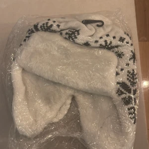 Vit och svart pälsfodrad trappermössa - Säljer en vit och svart trappermössa med stickat mönster och mjukt fuskpälsfoder. Mössan har öronlappar, flätade snören och en söt tofs på toppen. Perfekt för kalla vinterdagar när du vill hålla dig varm och ändå se snygg ut.