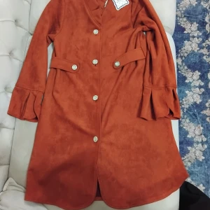 Rostbrun långklänning från Have Seeds - Snygg rostbrun långklänning från Have Seeds med dekorativa silverknappar framtill och coola volangdetaljer vid ärmsluten. Klänningen har lång ärm, markerad midja och är tillverkad i mjuk mockaliknande polyester. Perfekt för dig som gillar unika detaljer och statement-plagg.