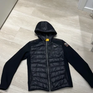 Svart pufferjacka från Parajumpers - Svart pufferjacka från Parajumpers med huva och hel dragkedja. Jackan har quiltad framsida och insida, samt en bröstficka med dragkedja. Ärmarna är släta och materialet är en mix av polyester och syntet. Klassisk patch på ena ärmen och orange detaljer på insidan.
