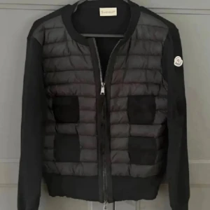 Svart pufferjacka från Moncler - Snygg svart pufferjacka från Moncler med quiltad front och stickade ärmar. Jackan har dragkedja framtill, två fickor och Moncler-logga på ena ärmen. Perfekt för dig som vill ha en stilren och exklusiv look.