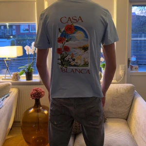 Ljusblå Casa Blanca t-shirt med tryck - Snygg ljusblå t-shirt från Casa Blanca med färgglatt tryck av blommor och landskap på ryggen samt mindre tryck på bröstet. T-shirten har rund hals och korta ärmar, perfekt för dig som gillar stilrena men ändå unika plagg.