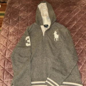 Grå hoodie från Polo Ralph Lauren L - Säljer en grå hoodie från Polo Ralph Lauren i storlek L. Tröjan har vit broderad logga på bröstet, en stor vit siffra 3 på ärmen och vita detaljer vid muddar och huva. Klassisk sportig look med huva och lång ärm, perfekt för chill dagar.