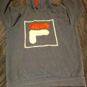 Mörkblå hoodie från FILA med logga - Snygg mörkblå hoodie från FILA i storlek M med stor vit och röd logga på bröstet. Tröjan har huva med röd insida och snörning, samt långa ärmar. Tillverkad i mjuk bomull som är skön att ha på sig. Perfekt för dig som gillar sportiga och stilrena plagg.