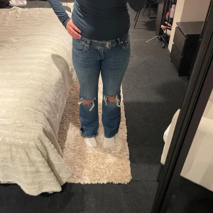 blå jeans med hål vid knäna  - Säljer ett par klassiska blå jeans med hål över knäna. Modellen har utsvängd passform och normal midja, med fem fickor och knappgylf. Jeansen är tillverkade i robust denim och har snygga detaljer vid benslutet.