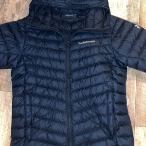 Mörkblå dunjacka Peak Performance - Snygg mörkblå dunjacka från Peak Performance med huva och dragkedja framtill. Jackan har quiltad design och logga på bröstet samt är tillverkad i lätt och slitstark polyester. Perfekt för kyliga dagar och har en clean look som funkar till det mesta.