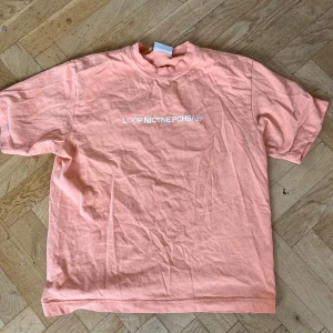 Persikofärgad t-shirt från Loop Nictne Pchs - Persikofärgad t-shirt med vit tryckt text 'LOOP NICTNE PCHS/ 18+' på bröstet. Klassisk rund hals och korta ärmar. Mjuk och skön bomullskvalitet, perfekt för chill dagar. Snyggt och enkelt statement-plagg för dig som gillar stilren design. Kan bara fås av säljare, sällsynt