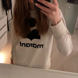 Vit sweatshirt från Isabel Marant - Säljer en vit sweatshirt från Isabel Marant med stort svart logotryck på bröstet. Tröjan har rund hals, långa ärmar och ribbade muddar vid ärmslut och nederkant. Perfekt för dig som gillar stilrena och trendiga plagg med statement.
