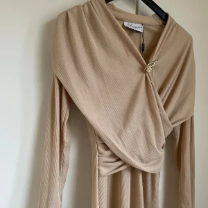 Beige omlottklänning från Closet  - Lång, ribbad beige klänning från Closet med omlott-detalj och elegant drapering framtill. Midilängd. Insydd i midjan. Brosch ingår ej