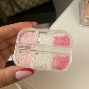 Nageldekorationer i rosa och vita blommor  - Söt ask med små nageldekorationer i form av blommor och pärlor i olika nyanser av rosa och vitt. Perfekt för att skapa kreativa och unika nageldesigner. Kommer i en praktisk plastförpackning med flera fack för enkel sortering.