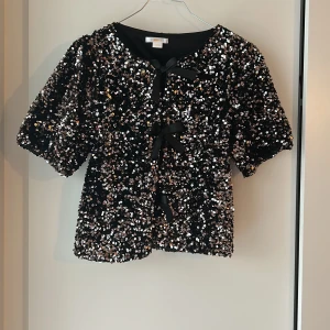 Svart paljettbolero från Gina Tricot - Glittrig svart bolerotopp från Gina Tricot, täckt med silvriga paljetter och tre snygga svarta rosettknyt framtill. Kortärmad och croppad modell som ger en extra festlig vibe. Perfekt för att sticka ut med coola detaljer och glans.